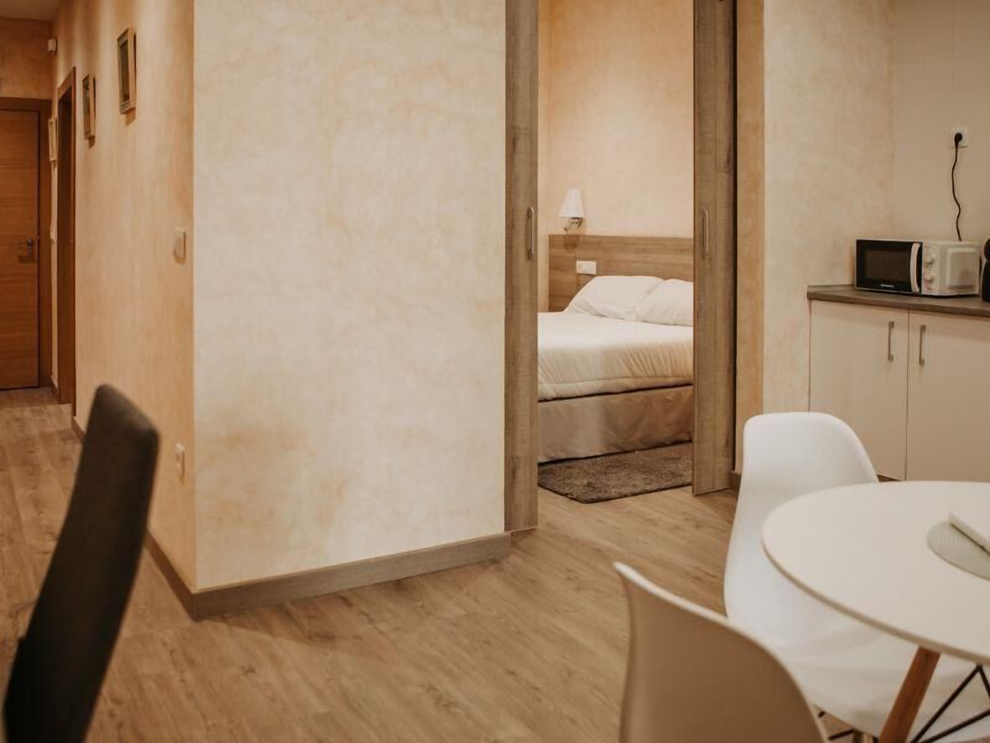 A modern hotel room with a wooden floor, a bed with white bedding, a small table with a chair, and a kitchenette with a microwave and kettle.
