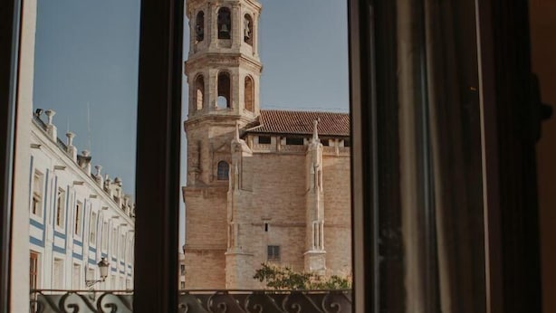 A view of a historic building with a bell tower and a clock, seen through a window with a decorative iron railing. - Apartamento 1 dormitorio y 2 sofas cama Atlántica (Valdepeñas)