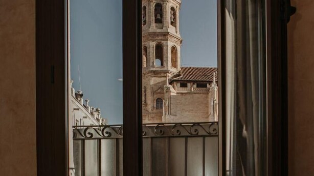 A view of a historic building with a clock tower through a window with a metal railing. - Apartamento Deluxe Mediterránea (Valdepeñas)