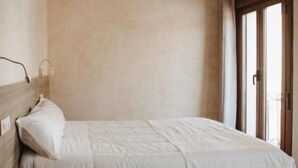 A neatly made bed with a wooden headboard, a bedside table, and a window with blinds. - Apartamento Deluxe Mediterránea (Valdepeñas)