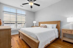 Une chambre à coucher avec un lit en bois, un ventilateur de plafond, une table de chevet, une lampe et une fenêtre avec des stores.