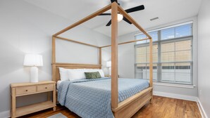 A bedroom with a four-poster bed, a nightstand, a bedside lamp, and a window with blinds.