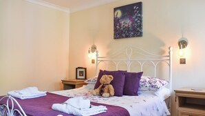 A neatly made bed with a purple bedspread, a white headboard, and a teddy bear on the pillows. There are two bedside tables with lamps, a framed picture on one, and a wooden nightstand with a lamp. - Gracies Cottage - UK44686 (Beamish)