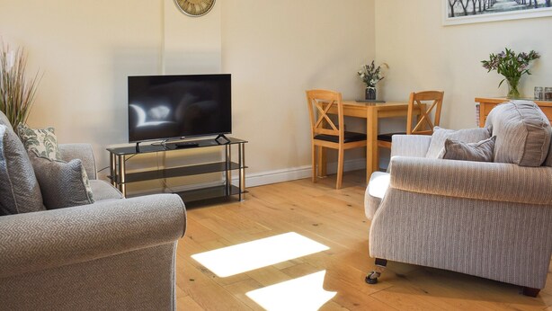 A living room with a flat-screen TV on a glass stand, wooden chairs, a sofa, and a dining table with a vase of flowers. - Gracies Cottage - UK44686 (Beamish)