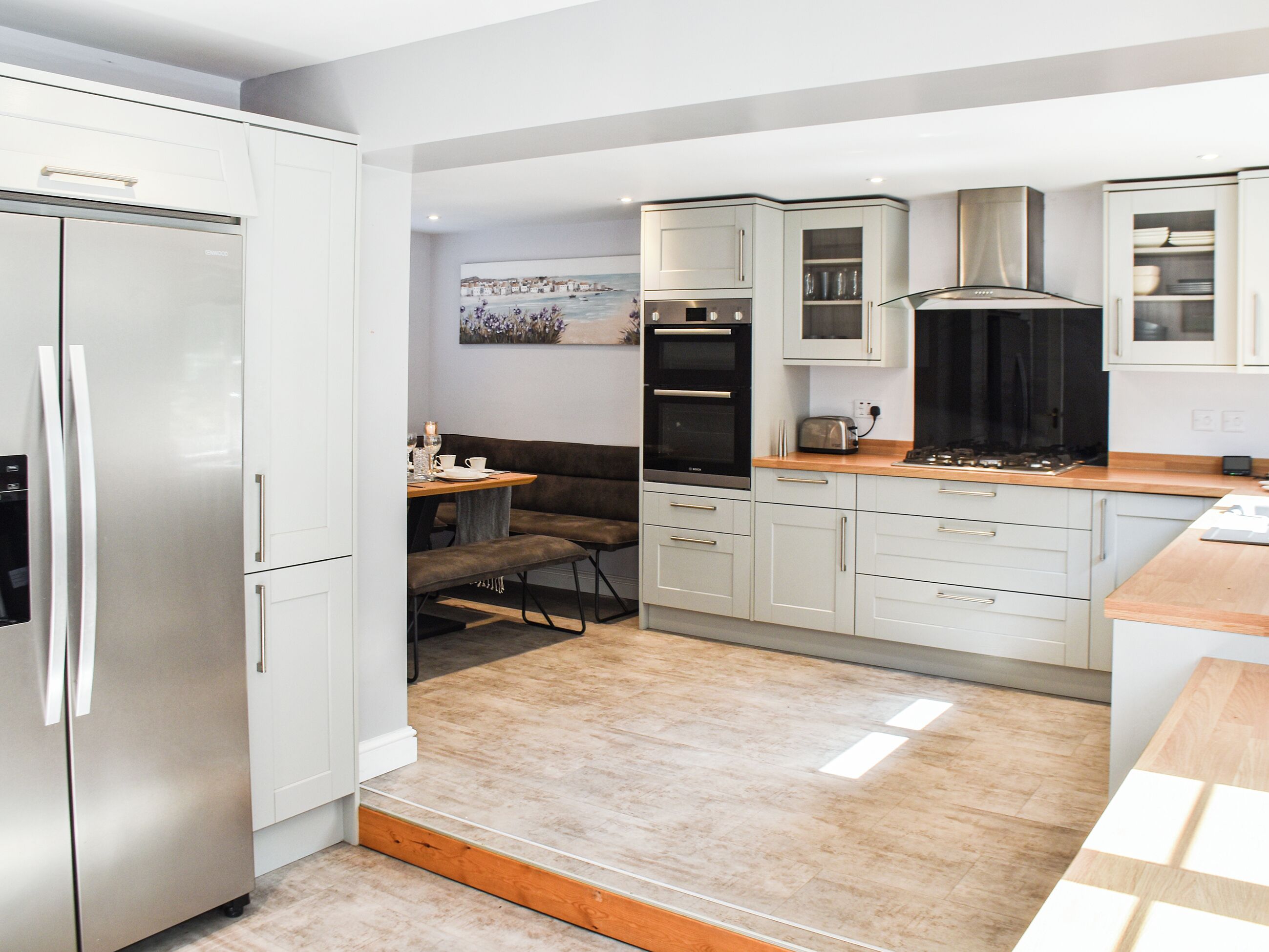 Une cuisine moderne avec des meubles blancs, un four encastré et un coin repas équipé d’une banquette.