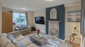 Ein modernes Wohnzimmer mit einer großen Couch, einem Couchtisch und einem Fernseher. Ein Kamin, ein Fenster mit Vorhängen und eine Pflanze auf einem Beistelltisch sind ebenfalls vorhanden.