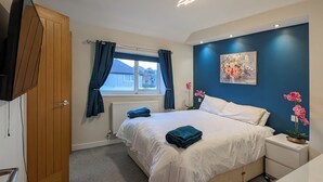 Ein Schlafzimmer mit Bett, Nachttisch, Fenster mit Vorhängen, Heizkörper, einem Bild an der Wand und einer Pflanze.