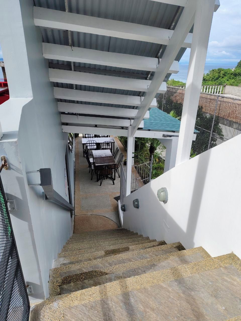 A staircase leading to a covered outdoor area with a table and chairs, surrounded by greenery and a view of the sea.