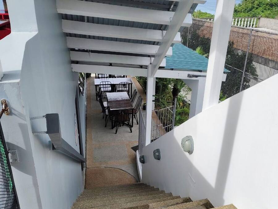A staircase leading to a covered outdoor area with a table and chairs, surrounded by greenery and a view of the sea.