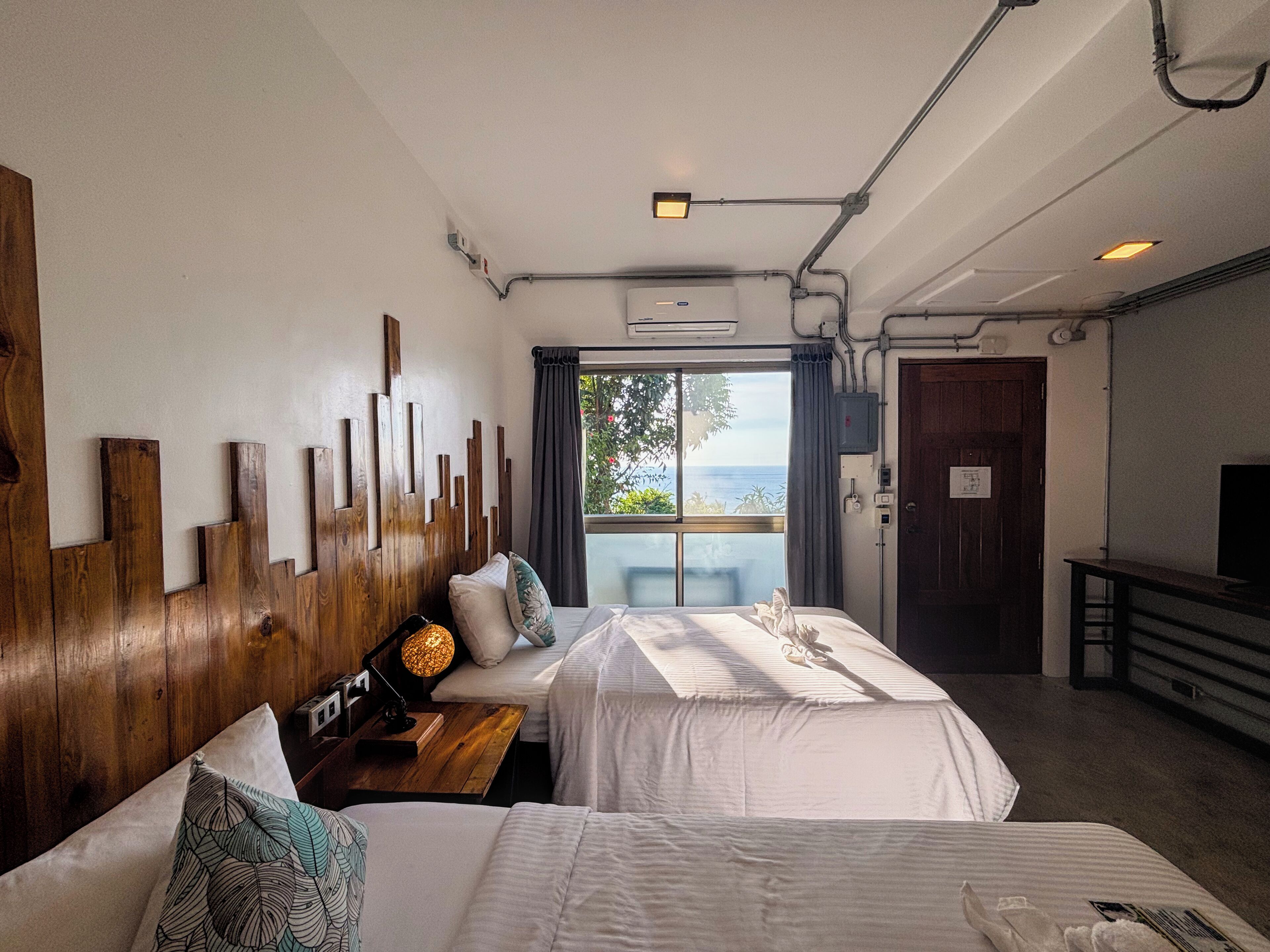 Habitación de hotel con dos camas, cabecero de madera, escritorio con lámpara, televisor y vista al mar a través de una ventana.