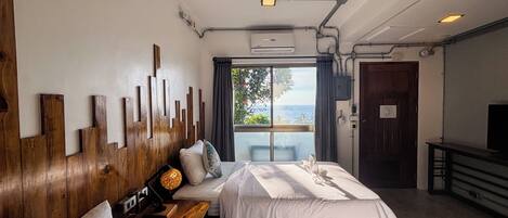 Habitación de hotel con dos camas, cabecero de madera, escritorio con lámpara, televisor y vista al mar a través de una ventana.
