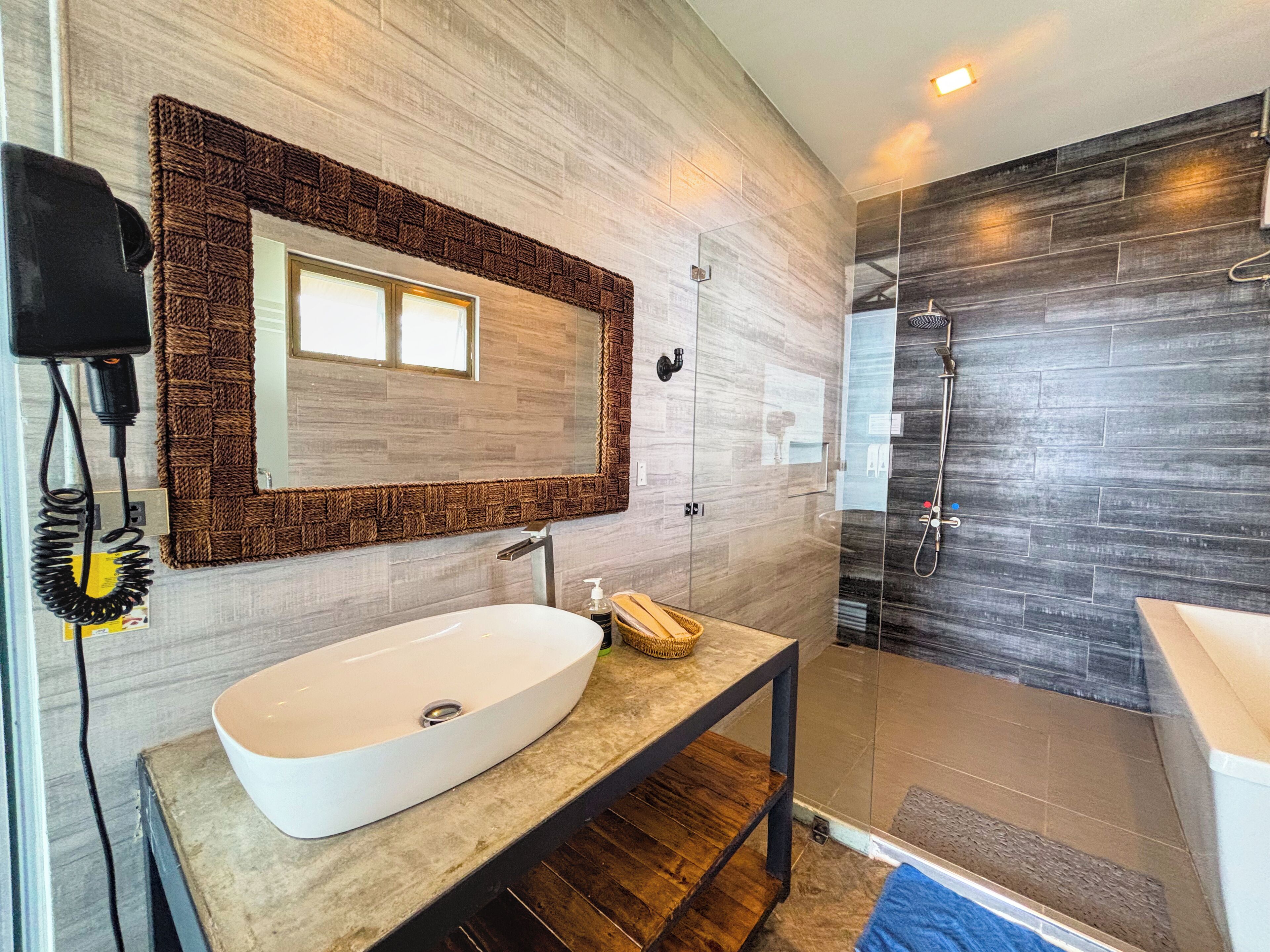 Un baño moderno con un lavamanos ovalado grande, un espejo con marco texturizado y una zona de ducha con azulejos oscuros.
