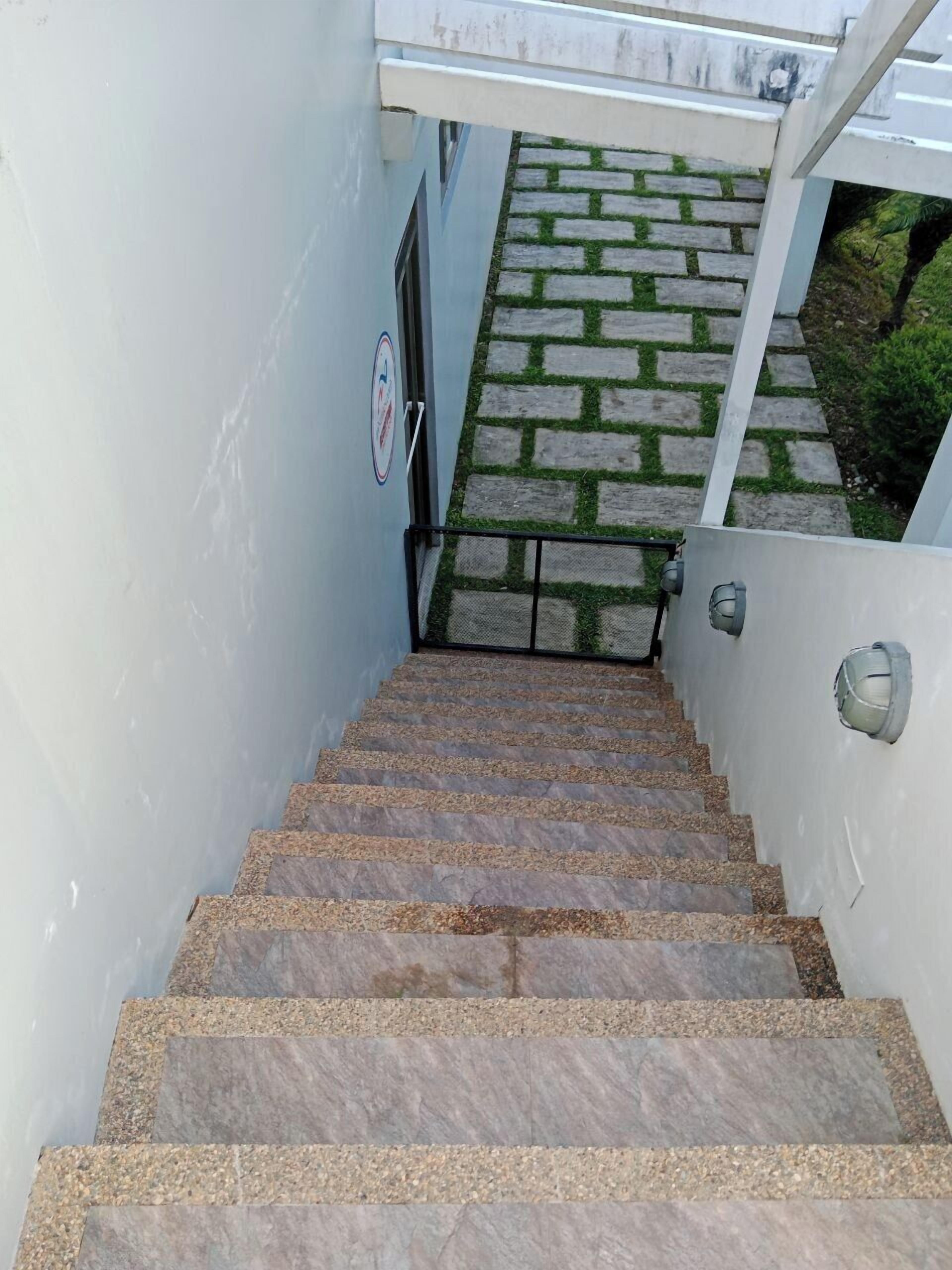 Una escalera de piedra conduce a una puerta, rodeada por un muro blanco y un sendero de piedra.