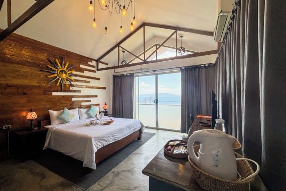 A bedroom with a bed, bedside table, lamp, and a view of the outdoors.