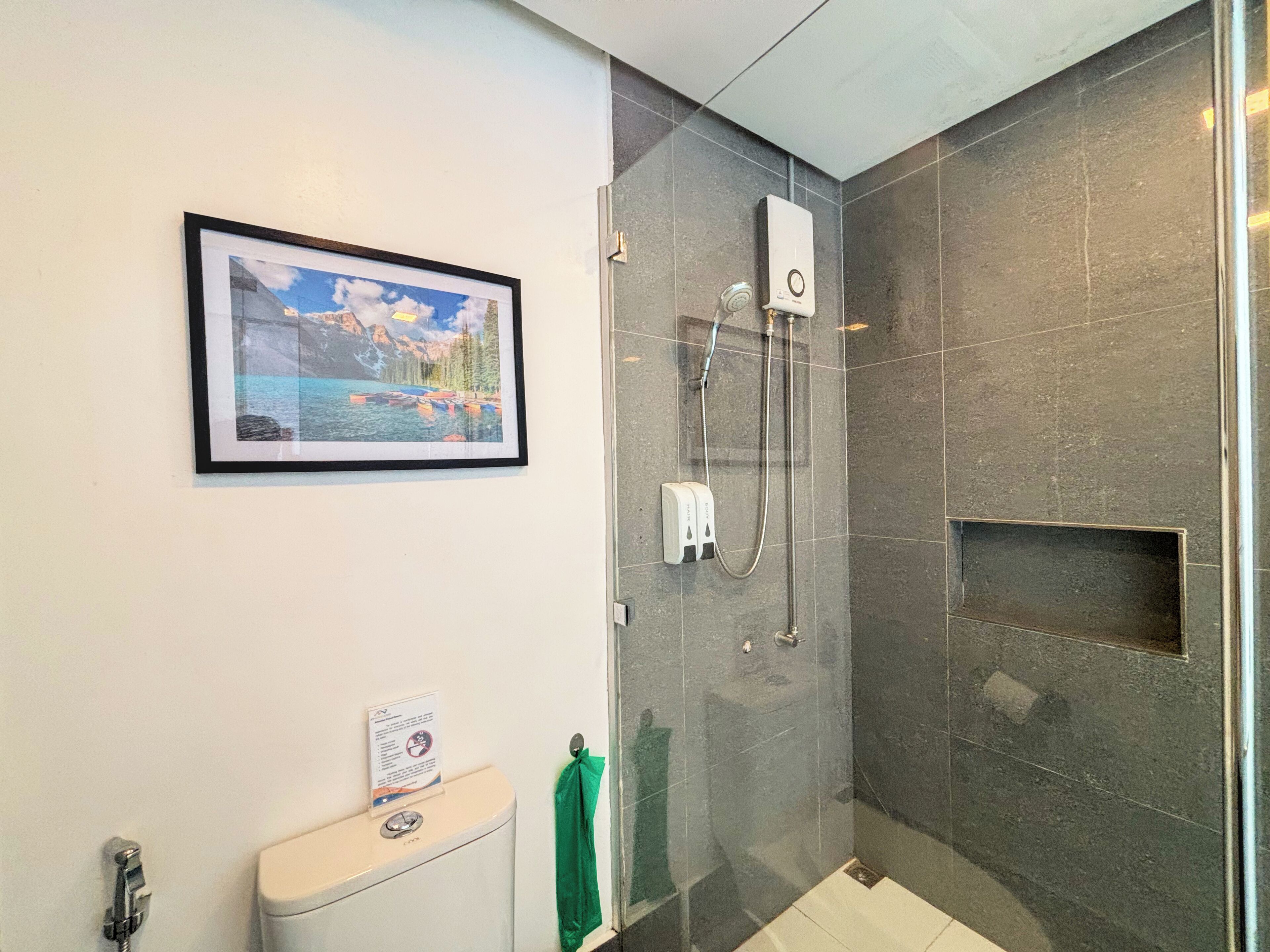 A modern bathroom with a glass-enclosed shower, a framed landscape picture on the wall, and a toilet with a green towel.