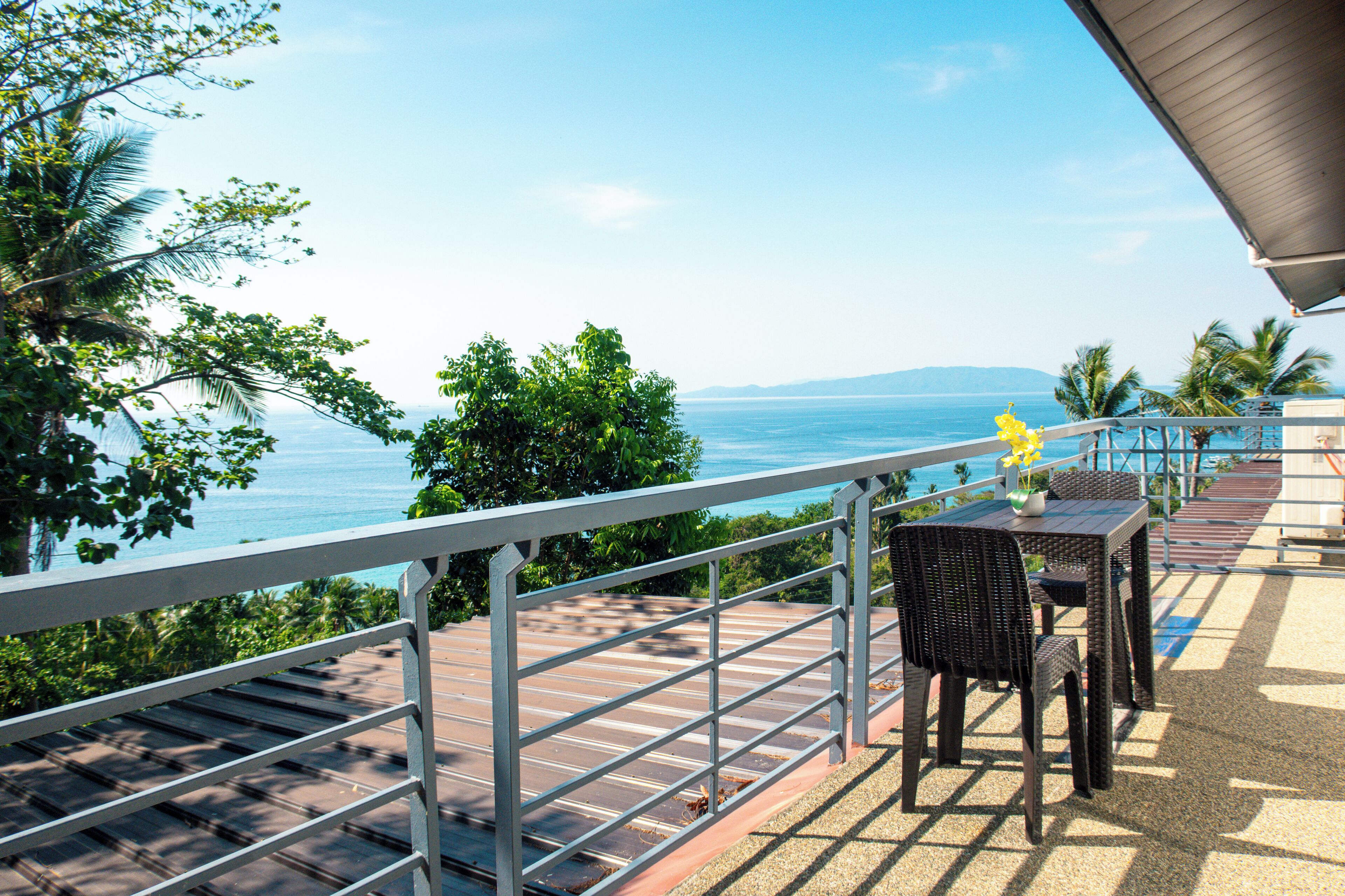 A terrace with a view of the sea, featuring a table and chairs, a railing, and tropical vegetation.