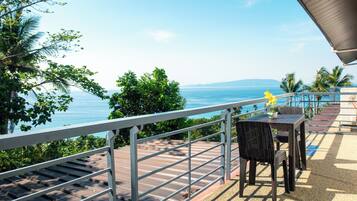 A terrace with a view of the sea, featuring a table and chairs, a railing, and tropical vegetation.