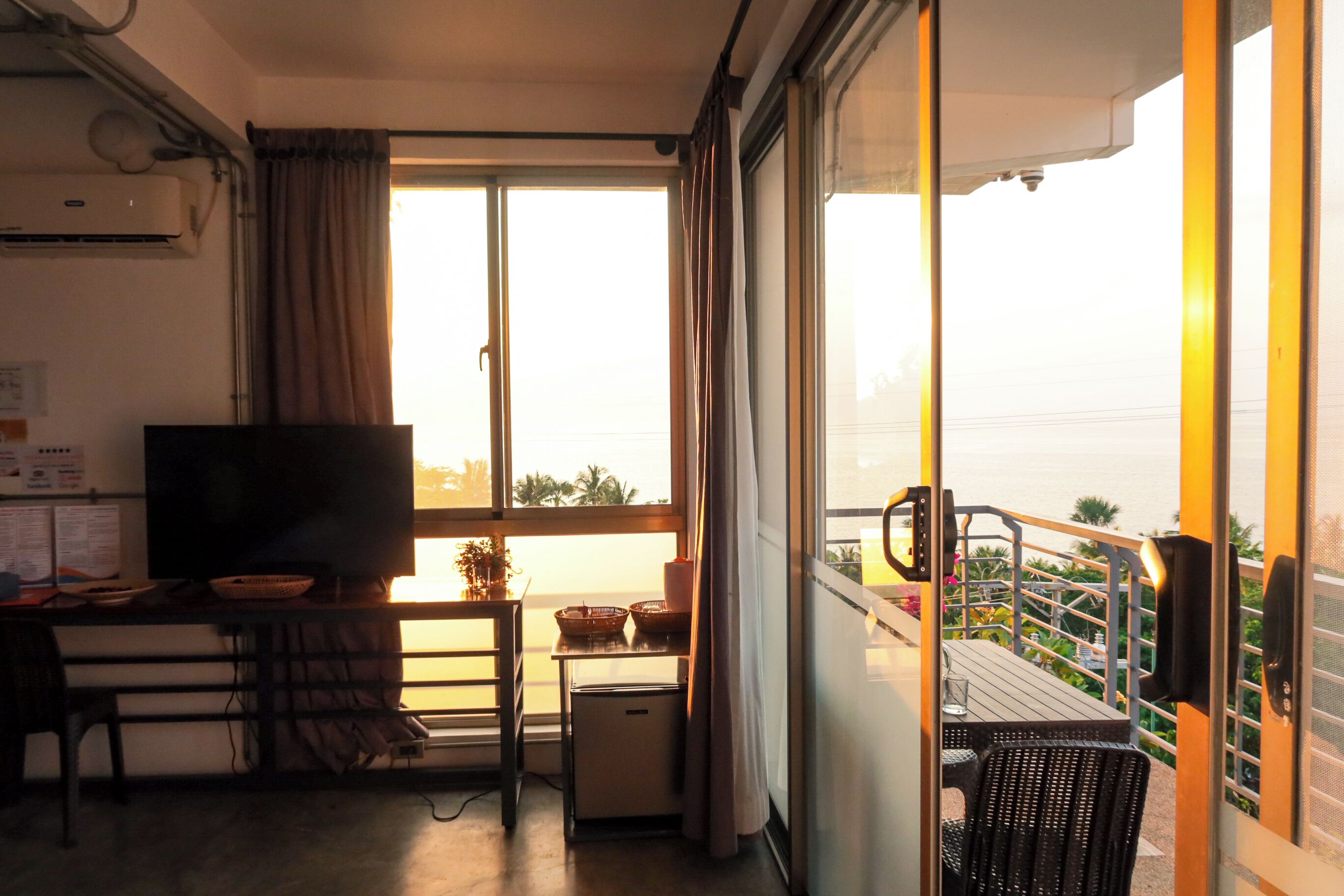 A balcony with a view of the sea, a TV, a desk with a chair, and a small refrigerator.