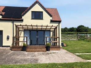 Un bâtiment à deux étages, doté d’un toit à deux versants, d’une entrée couverte avec une pergola en bois et d’un petit jardin agrémenté de plantes en pot.
