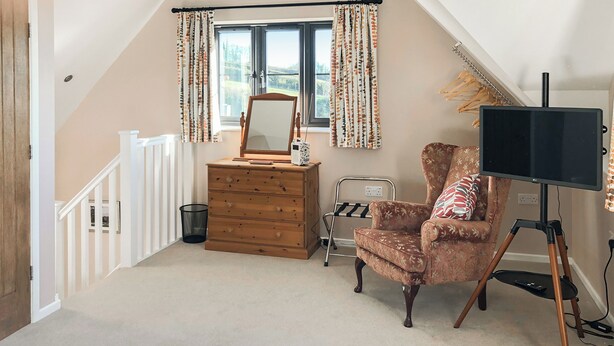 A bedroom with a wooden dresser, a chair, a mirror, a television on a tripod, and a window with curtains. - The Old Stable At Henley View (Draycott, near Cheddar)