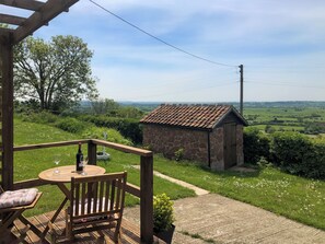 Une terrasse en bois avec une table et des chaises, un bâtiment en pierre et une vue sur la campagne.