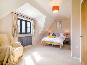 A bedroom with a sloped ceiling, a bed with white bedding, two bedside lamps, a radiator, and a window with curtains. - The Old Stable At Henley View (Draycott, near Cheddar)