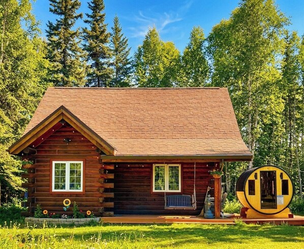 A log cabin with a pitched roof, a front porch, and a circular structure to the right.