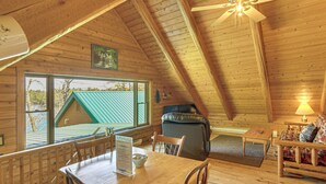 A wooden cabin interior with a dining table, chairs, a sofa, a ceiling fan, and a view of the outside.