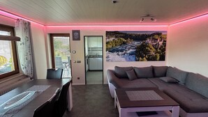 Uma sala de estar moderna com sofá, mesa de centro e área de jantar. Um grande quadro de paisagem está na parede, e o quarto tem teto de madeira.