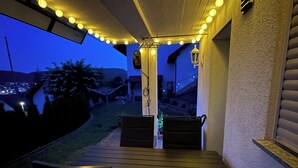 Um alpendre ao ar livre com mesa e cadeiras de madeira, luzes de corda e vista para o quintal.