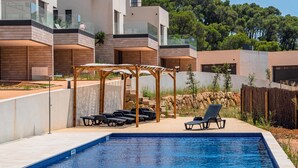 Une piscine extérieure moderne, dotée de chaises longues et d’une pergola, entourée d’un immeuble à plusieurs étages avec des balcons.