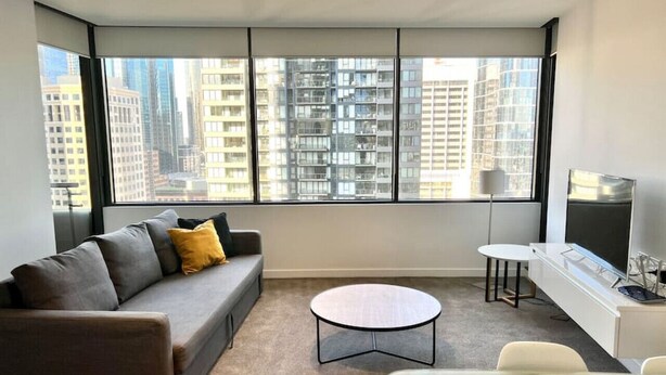 A modern living room with a grey sofa, a round coffee table, and a TV on a white cabinet. Large windows offer a view of tall buildings.