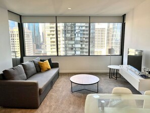 Un salon moderne avec un canapé gris, une table basse ronde et un téléviseur posé sur un meuble blanc. De grandes fenêtres offrent une vue sur de hauts immebles.