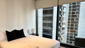 A modern hotel room with a large bed, a nightstand, a lamp, and a view of a high-rise building through a large window.