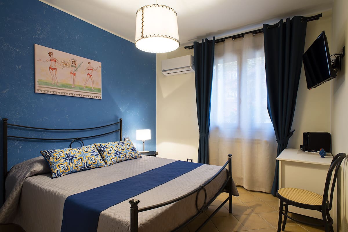 A bedroom with a bed, a desk, a chair, a television, and a painting on the wall.