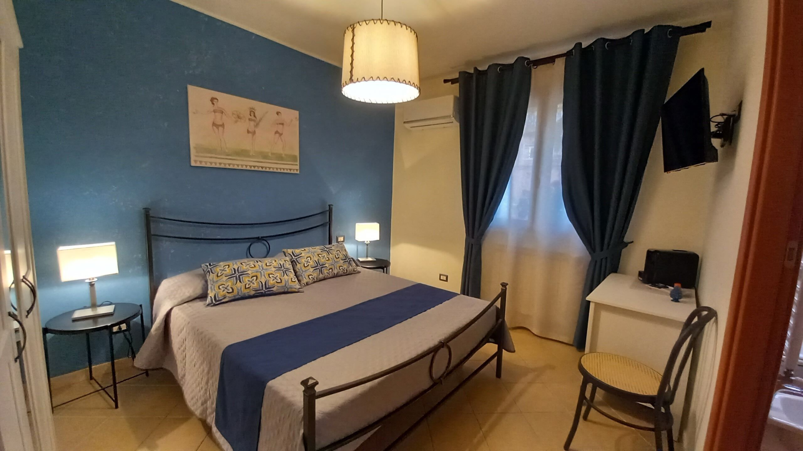 Ein Schlafzimmer mit einem Bett, zwei Nachttischen mit Lampen, einem Schreibtisch mit Stuhl, einem Wand-Fernseher und einem Fenster mit Vorhängen.