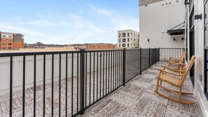 Un balcón con barandilla de metal negro, sillas mecedoras de madera y vista a edificios al fondo.