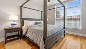 Una cama con dosel de madera y colcha gris, una mesita de noche con lámpara, y vistas a un paisaje urbano a través de amplios ventanales.