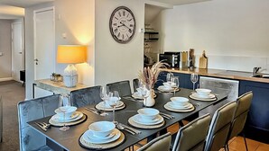 Uma sala de jantar com mesa posta para seis, cadeiras, um relógio e cozinha ao fundo.