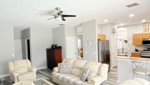 A living room with a sofa set, a coffee table, and a lamp. There is a ceiling fan and a kitchen area in the background. - High Grove Hideaway, Spacious 5BR Villa w/ Pool Near Disney (Four Corners)