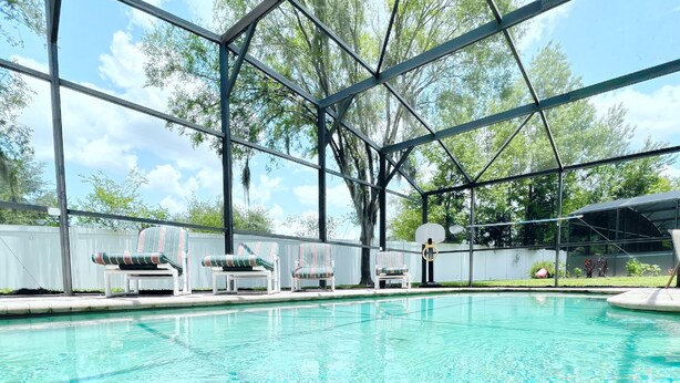 A clear swimming pool with a metal roof structure, surrounded by lounge chairs and a white fence. - High Grove Hideaway, Spacious 5BR Villa w/ Pool Near Disney (Four Corners)
