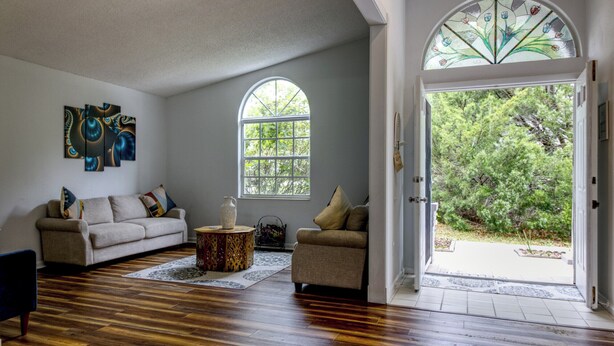 A living room with hardwood flooring, a beige sofa, a round coffee table, and a large window with a view of greenery.