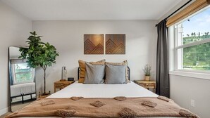 Un dormitorio con una cama grande, dos mesitas de noche, un espejo, una planta en maceta y una ventana con cortinas.