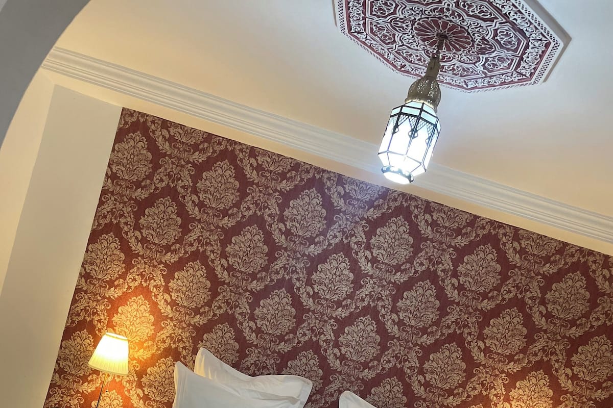 A neatly made bed with white linens and two pillows, a patterned wallpaper in shades of red and brown, and two bedside lamps on either side.