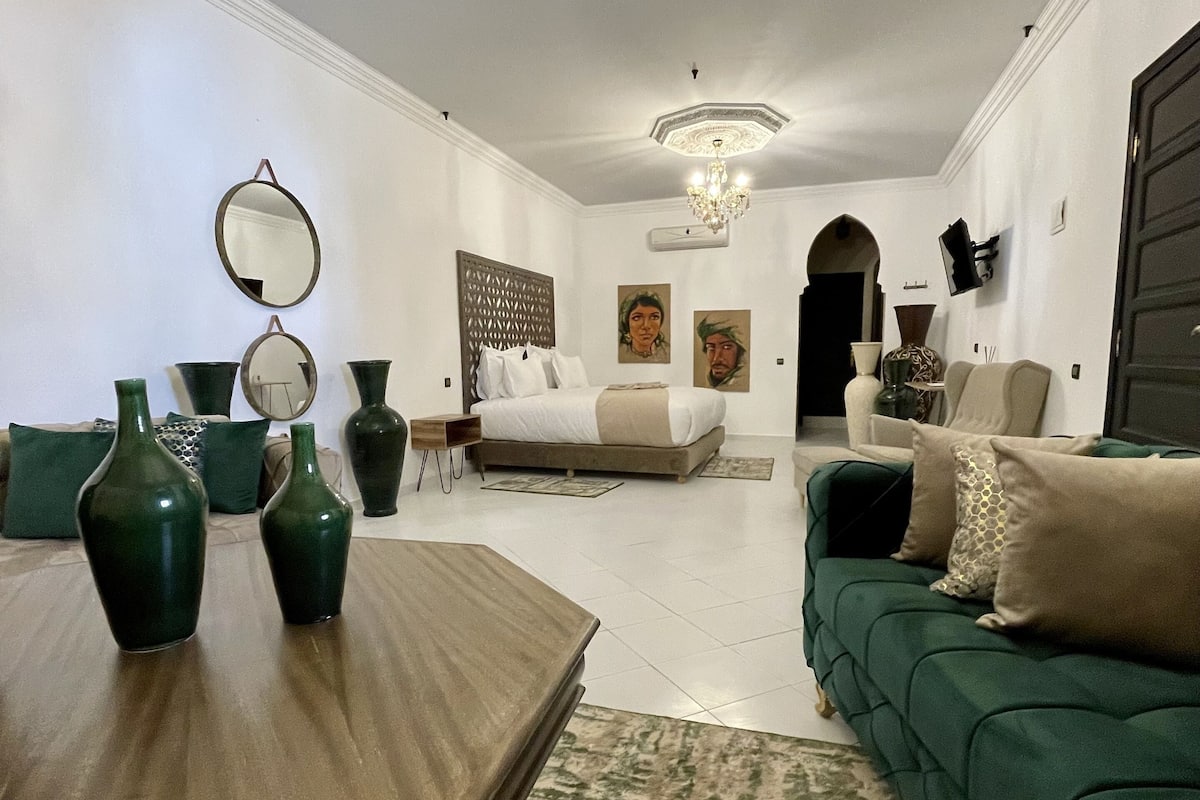 A spacious living room with a large sofa, a wooden coffee table, and two green vases. There is a chandelier hanging from the ceiling and a large mirror on the wall.