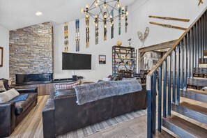 Un salon moderne avec un mur en pierre, un grand téléviseur à écran plat, un canapé en cuir foncé et un escalier avec une rampe en bois.