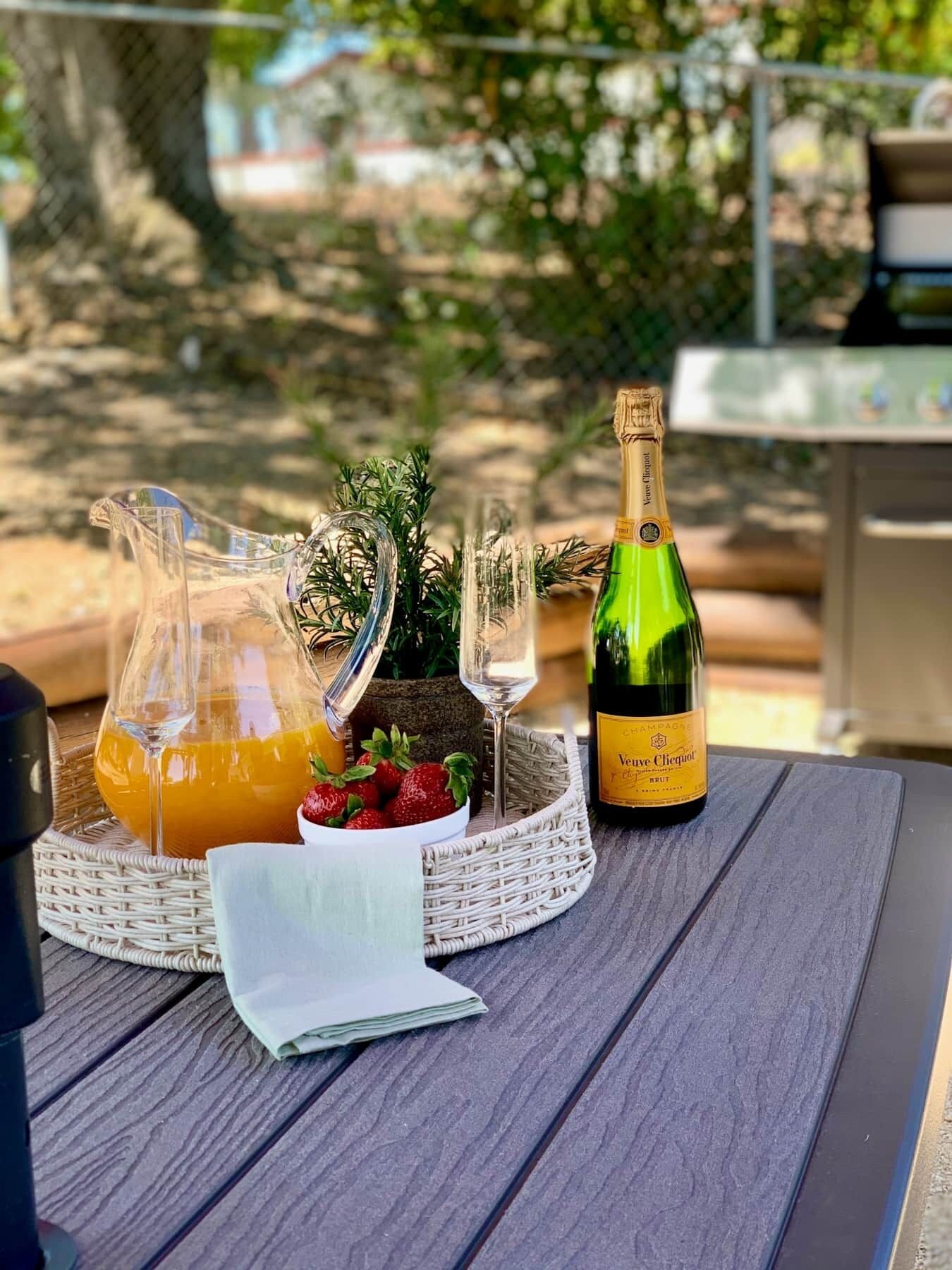 Une table dressée avec une bouteille de champagne, un pichet de jus d’orange, un verre et un panier rempli de fraises et de verdure.