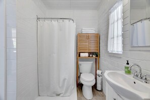 A compact bathroom with white tiled walls, a shower curtain, a toilet, a wooden cabinet with towels, and a sink with a mirror. - Hilltop Paso: Dog Friendly, Firepit, BBQ, Views (Paso Robles)