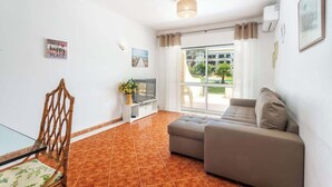 Ein Wohnzimmer mit einer Couch, einem Fernseher, einem Sessel und einem Fenster mit Blick auf Gebäude und Grünflächen.