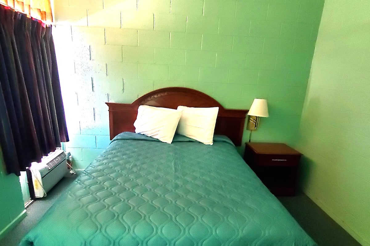 A single bed with a green quilt, two pillows, a wooden headboard, a nightstand with a lamp, and a wall-mounted heater.